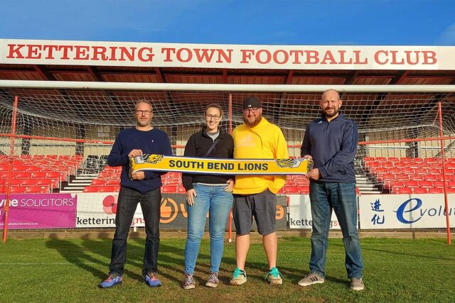 Ritchie, Sarah, Ryan, and Chris hold up a South Bend Lions flag at Kettering Town Football Club